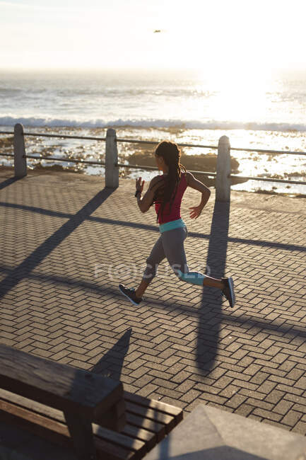 Mujer afroamericana haciendo ejercicio en un paseo marítimo corriendo. Fitness estilo de vida saludable al aire libre. - foto de stock