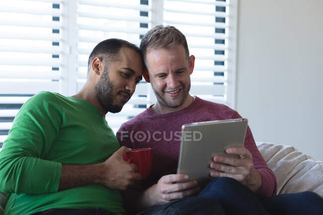 Casal masculino gay multi étnico sorrindo e sentado no sofá bebendo café e usando tablet em casa. Ficar em casa em auto-isolamento durante o bloqueio de quarentena. — Fotografia de Stock