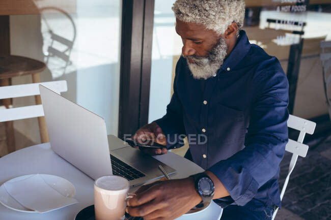 Ein afroamerikanischer Senior sitzt mit Laptop und Smartphone am Tisch vor einem Café. digitaler Nomade in der Stadt unterwegs. — Stockfoto