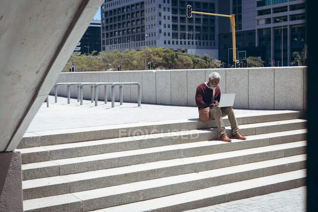 Homme âgé afro-américain assis sur les marches dans la rue à l'aide d'un ordinateur portable. nomade numérique dans la ville. — Photo de stock