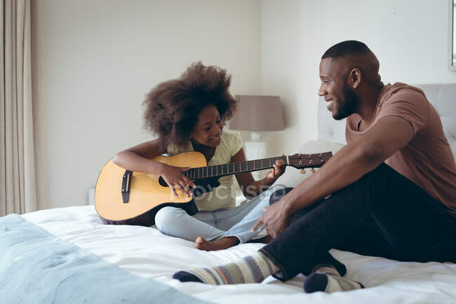 Hombre afroamericano y su hija sentado en la cama chica está tocando la guitarra. permanecer en casa en aislamiento durante el bloqueo de cuarentena. - foto de stock