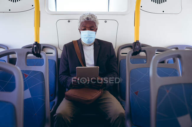 Hombre mayor afroamericano con máscara facial sentado en el autobús usando tableta digital. nómada digital en la ciudad durante la pandemia de coronavirus covid 19. - foto de stock