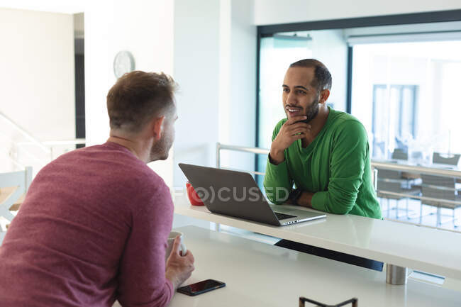 Multi ethnic gay male couple smiling, sitting in kitchen drinking coffee and using laptop at home. Staying at home in self isolation during quarantine lockdown. — Stock Photo
