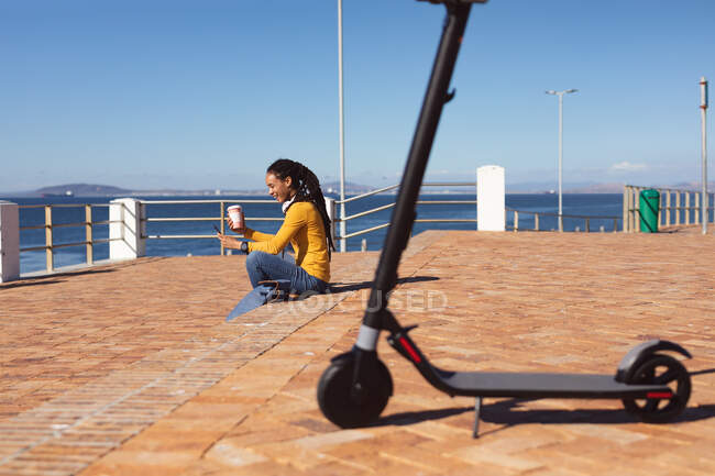 Smiling african american woman wearing headphones sitting using smartphone on promenade by the sea. Digital nomad on the go lifestyle. — Stock Photo