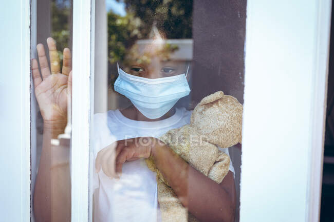 Chica afroamericana con máscara facial sosteniendo oso de peluche saludando por la ventana. permanecer en casa en aislamiento durante el bloqueo de cuarentena. - foto de stock