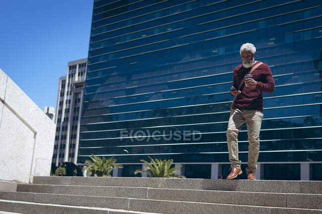 Hombre mayor afroamericano caminando por los escalones fuera del edificio sosteniendo café para llevar y usando un teléfono inteligente. nómada digital en la ciudad. - foto de stock