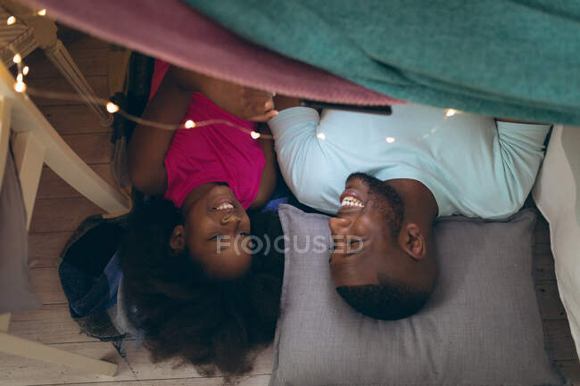 Niña afroamericana y su padre yaciendo en un fuerte. permanecer en casa en aislamiento durante el bloqueo de cuarentena. - foto de stock