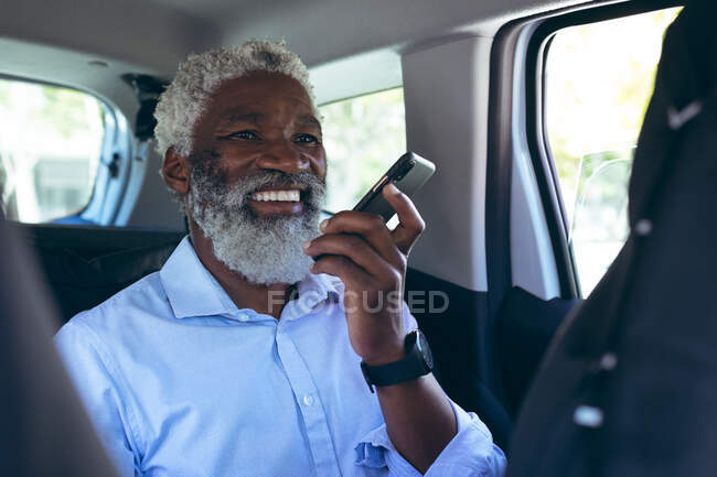 Homme âgé afro-américain assis dans un taxi parlant sur smartphone et souriant. nomade numérique dans la ville. — Photo de stock