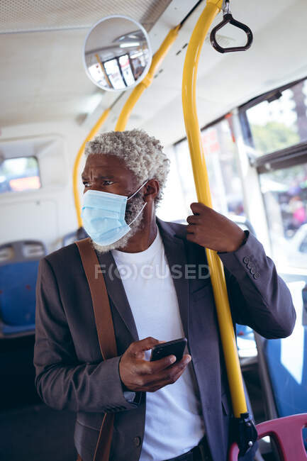 Ein afroamerikanischer älterer Mann mit Mundschutz steht im Bus und hält sein Smartphone in der Hand. digitaler Nomade unterwegs in der Stadt während Coronavirus covid 19 Pandemie. — Stockfoto