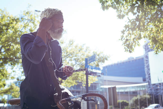 Ein afroamerikanischer Senior sitzt auf einem Fahrrad in der Straße und setzt sich Kopfhörer auf. digitaler Nomade in der Stadt unterwegs. — Stockfoto
