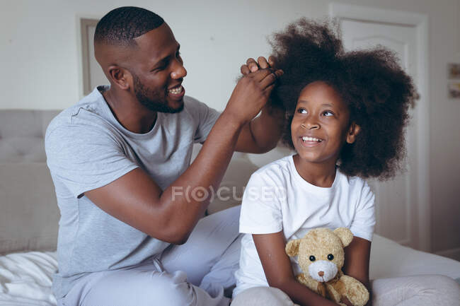 Uomo afroamericano seduto sul letto a fare i capelli alla figlia. stare a casa in isolamento durante l'isolamento in quarantena. — Foto stock