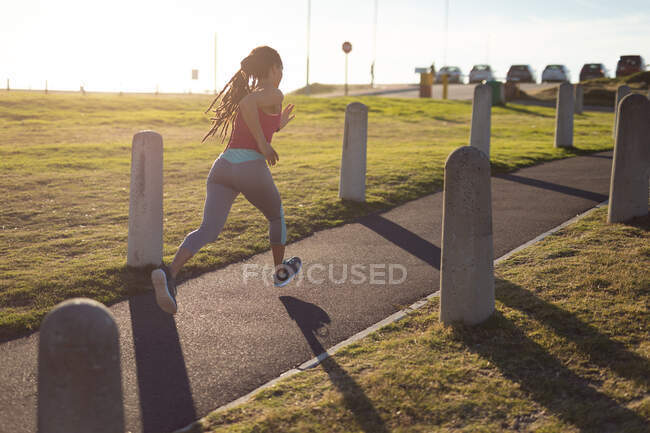 Mujer afroamericana haciendo ejercicio en el parque, corriendo por el camino en un día soleado. Fitness estilo de vida saludable al aire libre. - foto de stock