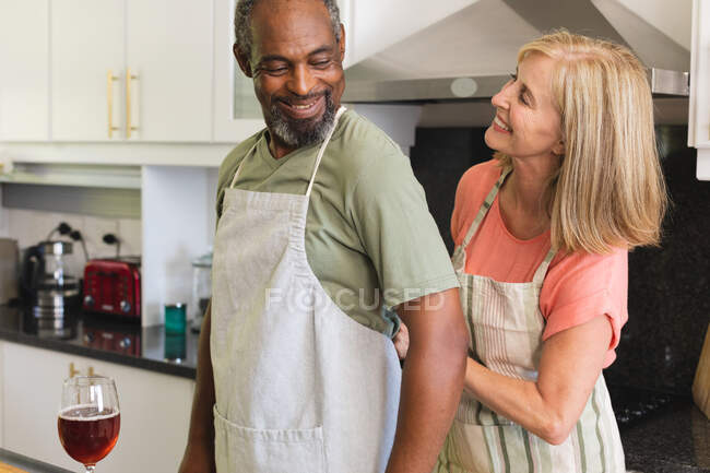 Casal sênior diverso que põe aventais na cozinha antes de cozinhar. ficar em casa em isolamento durante o confinamento de quarentena. — Fotografia de Stock