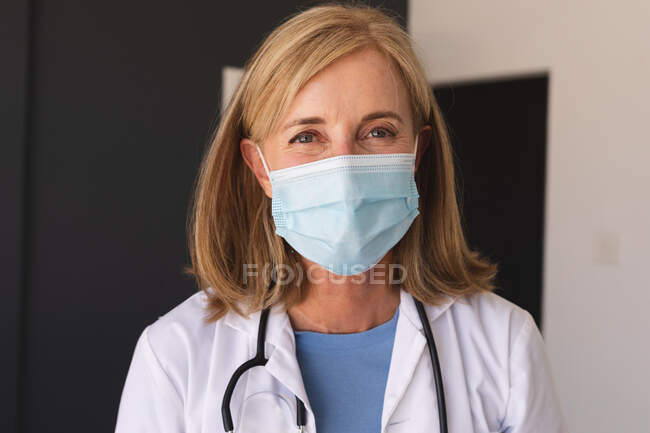 Doctora mayor caucásica vistiendo una máscara facial mirando a la cámara y sonriendo. profesional médico en el trabajo durante la pandemia del coronavirus covid 19. - foto de stock