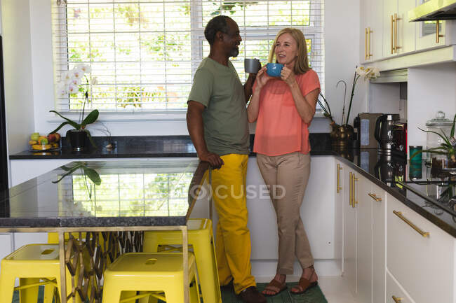 Разнообразная старшая пара, стоящая на кухне и пьющая кофе. оставаться дома в изоляции во время карантинной изоляции. — стоковое фото