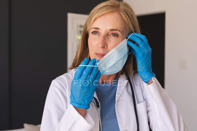 Doctora mayor caucásica poniéndose una mascarilla. profesional médico en el trabajo durante la pandemia del coronavirus covid 19. - foto de stock
