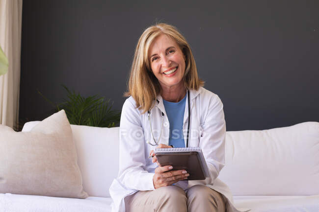 Caucasiana médico sênior do sexo feminino sentado no sofá sorrindo dando videochamada consulta. telemedicina em casa durante o bloqueio de quarentena. — Fotografia de Stock