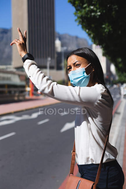 Gemischte Frau mit Gesichtsmaske, die auf der Straße steht und ein Taxi begrüßt. Frau während Coronavirus-Pandemie in der Stadt unterwegs. — Stockfoto