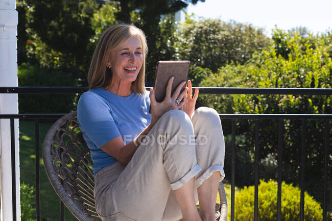 Mulher idosa caucasiana sentada no terraço usando tablet digital e sorrindo. ficar em casa em isolamento durante o confinamento de quarentena. — Fotografia de Stock