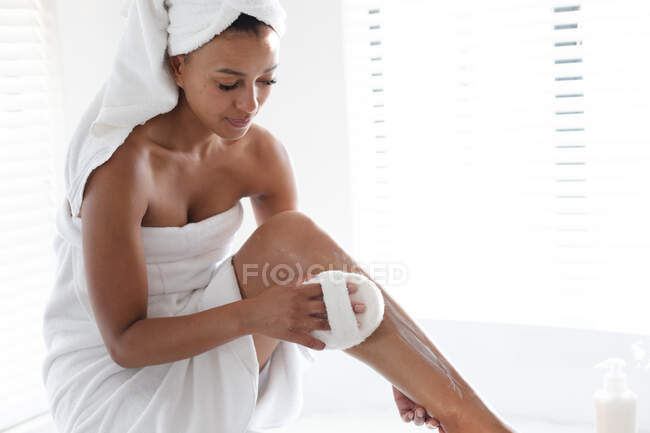 African american woman massaging her legs with loofah pad in bathroom. staying at home in self isolation in quarantine lockdown — Stock Photo