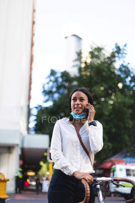 Mujer de raza mixta con máscara facial hablando en smartphone caminando por la calle. mujer en la salida y alrededor de la ciudad durante coronavirus covid 19 pandemia. - foto de stock