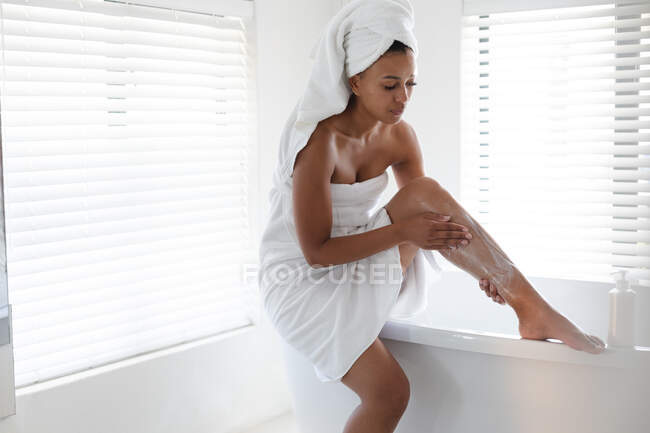Mujer afroamericana aplicando crema hidratante en sus piernas en el baño. permaneciendo en casa en aislamiento en cuarentena - foto de stock