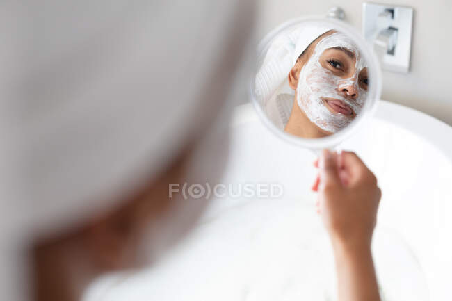 Африканская американка в маске для лица держит зеркало в ванной. оставаться дома в изоляции в карантинной изоляции — стоковое фото