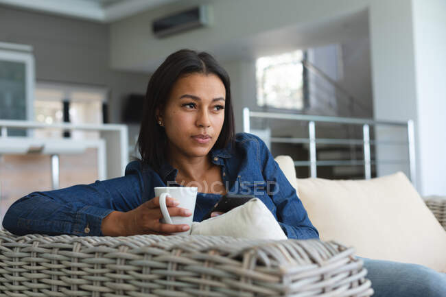 Femme de course mixte se détendre dans le salon sur le canapé avec une tasse de café. rester à la maison dans l'isolement pendant le confinement en quarantaine. — Photo de stock