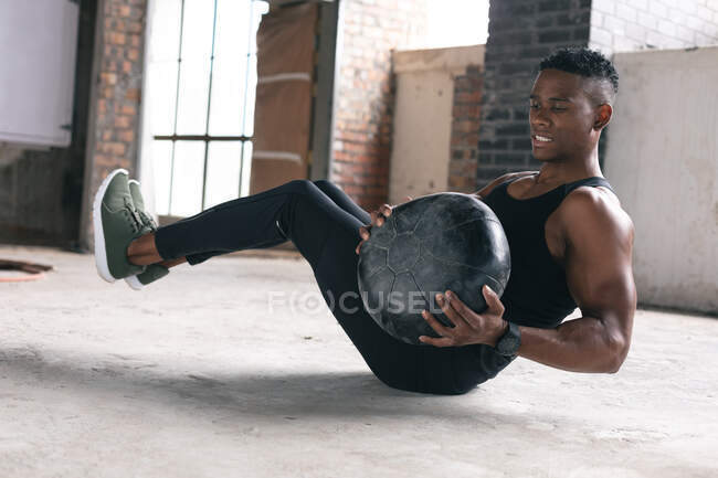 Afro-americano exercitando-se no armazém fazendo sit ups segurando bola medicina. estilo de vida urbano ativo saudável. — Fotografia de Stock