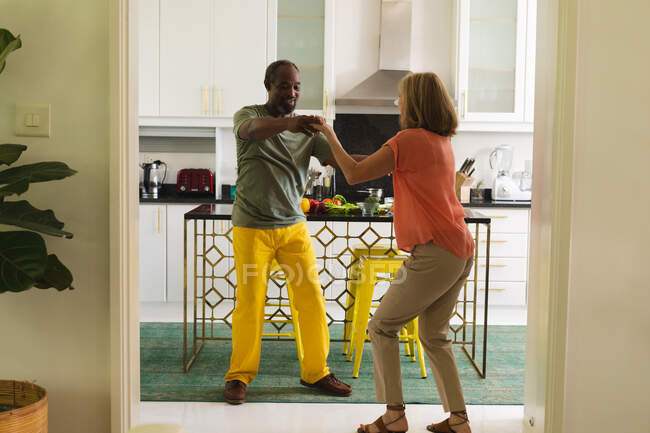 Casal sênior diverso dançando na cozinha e sorrindo. ficar em casa em isolamento durante o confinamento de quarentena. — Fotografia de Stock