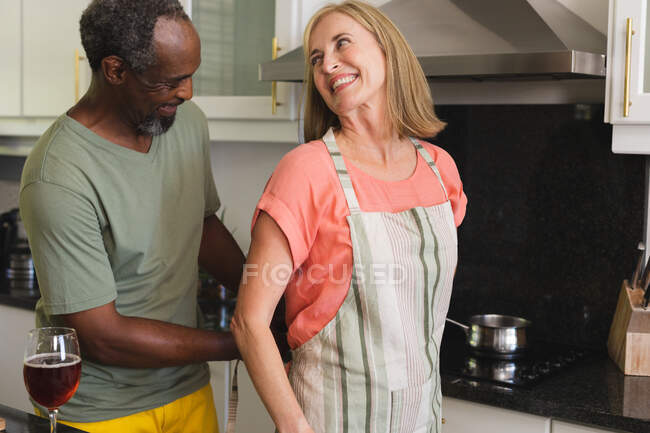 Diverse senior couple putting on aprons in kitchen before cooking. staying at home in isolation during quarantine lockdown. — Stock Photo