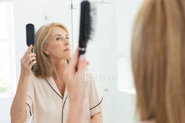 Mulher idosa caucasiana de pé na casa de banho a escovar o cabelo. ficar em casa em isolamento durante o confinamento de quarentena. — Fotografia de Stock