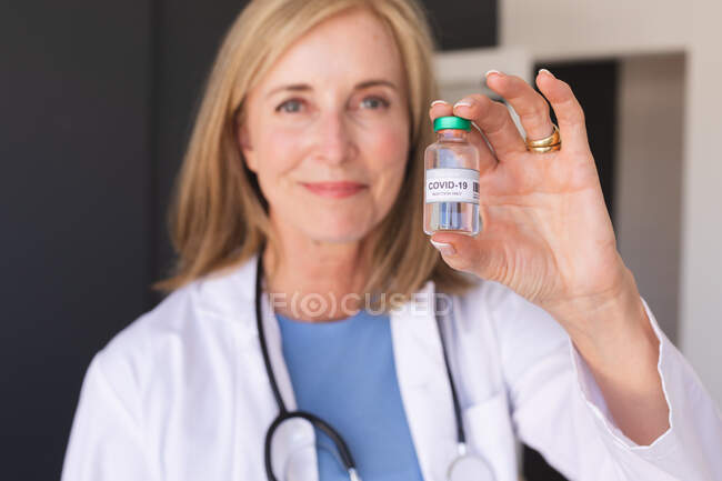 Doctora mayor caucásica sosteniendo la vacuna covid-19 y sonriendo. profesional médico en el trabajo durante la pandemia del coronavirus covid 19. - foto de stock