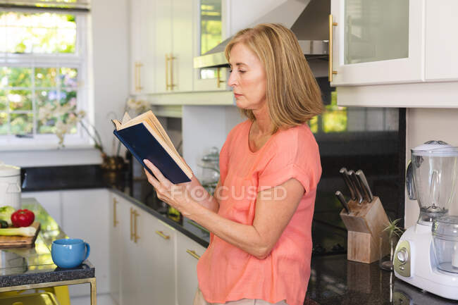 Старшая белая женщина, сидящая на кухне и читающая книгу. оставаться дома в изоляции во время карантинной изоляции. — стоковое фото