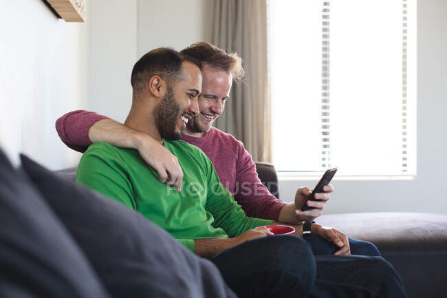 Мультинациональная гей-пара улыбается, сидит на диване и использует смартфон дома. оставаться дома в изоляции во время карантинной изоляции. — стоковое фото
