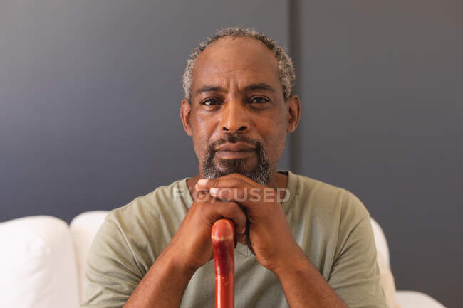 Portrait of mixed race senior man sitting on couch holding cane looking at camera and smiling. staying at home in isolation during quarantine lockdown. — Stock Photo