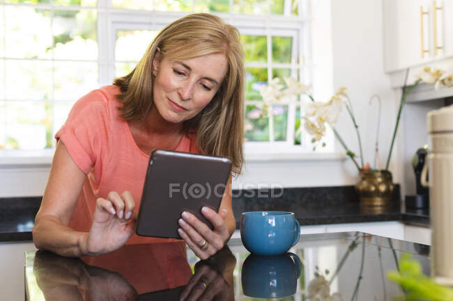 Старшая белая женщина, сидящая на кухне с цифровым планшетом. оставаться дома в изоляции во время карантинной изоляции. — стоковое фото