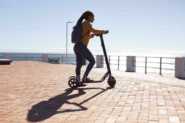 Sorrindo mulher afro-americana admirando a vista do mar com scooter no passeio marítimo. nômade digital em movimento estilo de vida. — Fotografia de Stock