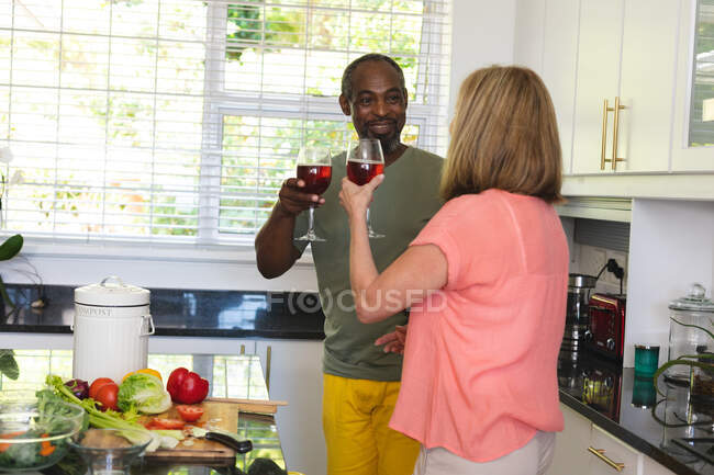 Diversa pareja de ancianos de pie en la cocina haciendo un brindis con copas de vino. permanecer en casa aislado durante el bloqueo de cuarentena. - foto de stock