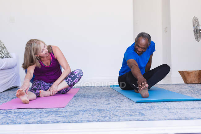 Diverso casal sênior exercitando sentado em tapetes de ioga alongamento. ficar em casa em isolamento durante o confinamento de quarentena. — Fotografia de Stock
