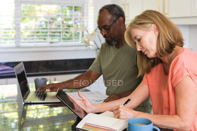 Разнообразная старшая пара, сидящая на кухне с ноутбуком и просматривающая документы. оставаться дома в изоляции во время карантинной изоляции. — стоковое фото
