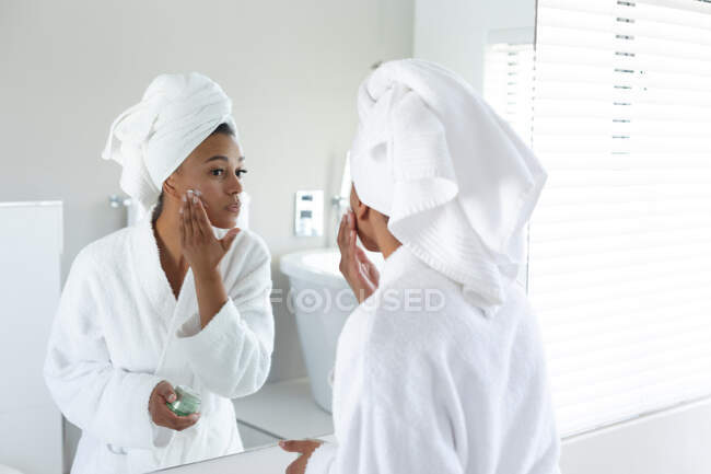 Африканська американка у халаті для ванни, дивлячись у дзеркало у ванній кімнаті, застосовує крем для обличчя. Залишатися вдома в ізоляції в карантині. — стокове фото