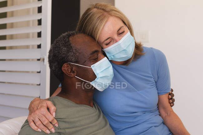 Diverso casal sênior usando máscaras faciais sentado no sofá e abraçando. ficar em casa em isolamento durante o confinamento de quarentena. — Fotografia de Stock
