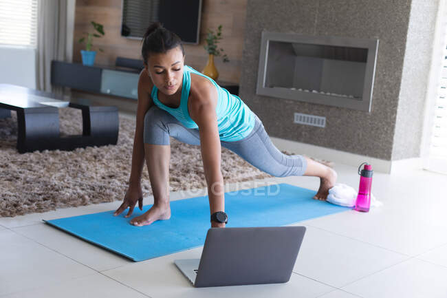 Mujer afroamericana practicando yoga mientras mira la pantalla del portátil en casa. permaneciendo en casa en aislamiento en cuarentena - foto de stock