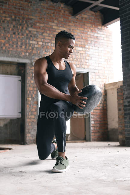 African american man exercising in warehouse doing lunges holding medicine ball. healthy active urban lifestyle. — Stock Photo