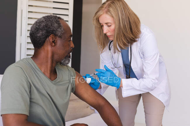 Médica idosa caucasiana a vacinar um homem de raça mista. profissional médico a trabalhar durante a pandemia do coronavírus covid 19. — Fotografia de Stock