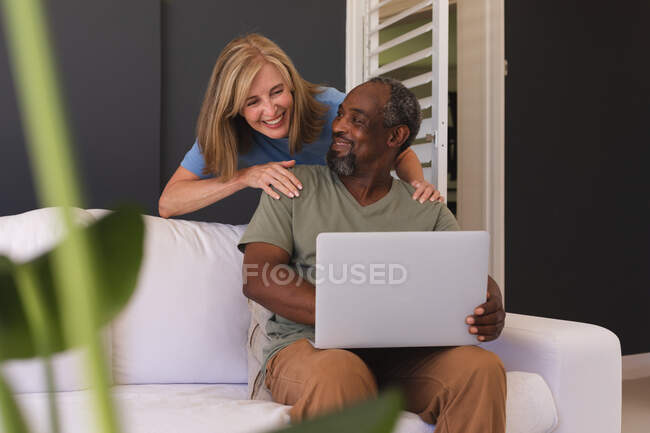 Diverse senior couple sitting on couch and using laptop. staying at home in isolation during quarantine lockdown. — Stock Photo