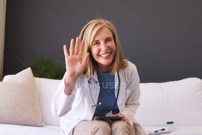 Medico donna anziana caucasica seduta sul divano sorridente dando consultazione videochiamata. telemedicina a casa durante la quarantena. — Foto stock