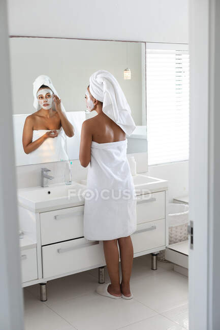 Африканская американка надевает маску для лица, глядя в зеркало ванной комнаты. оставаться дома в изоляции в карантинной изоляции — стоковое фото