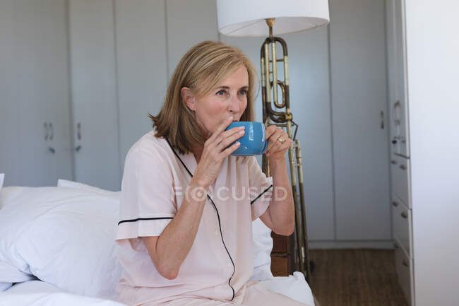 Кавказька старша жінка сидить на ліжку і п'є чашку кави. Перебуваючи вдома в ізоляції під час карантину.. — стокове фото